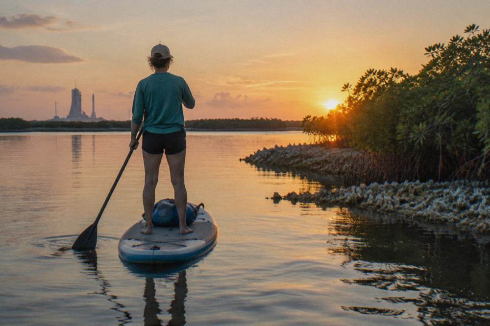 A paddleboarder standing on calm lagoon water as the sun sets behind mangroves