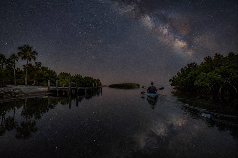 Single kayak gliding across mirror-calm lagoon water under a dark star-filled sky
