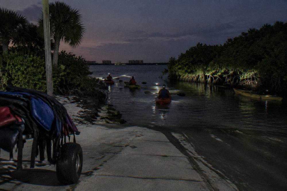 Group of kayaks with headlamps gathered at a mangrove-lined launch ramp before a night paddle
