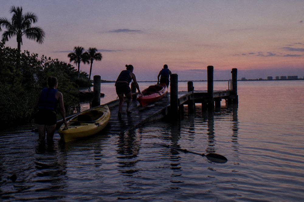 Paddlers in life jackets launching kayaks from a dark shoreline at dusk
