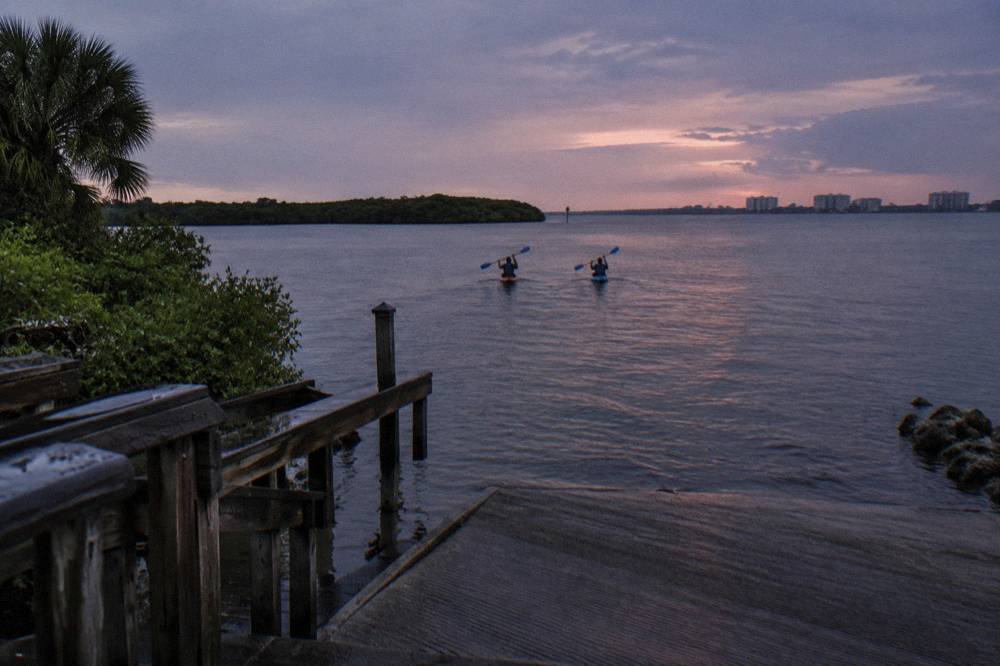 Two kayakers on a dark lagoon at dusk with paddles lifted and faint blue light trailing from the blades