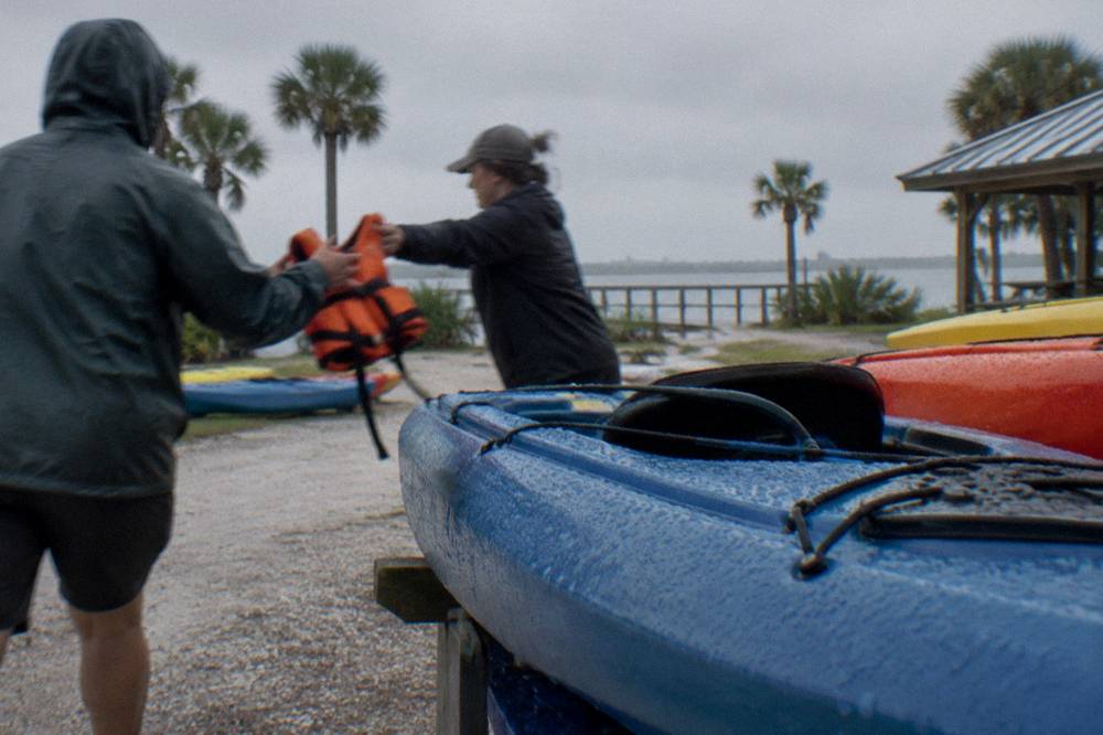 A guide handing a life jacket to a guest beside stacked kayaks at a launch site