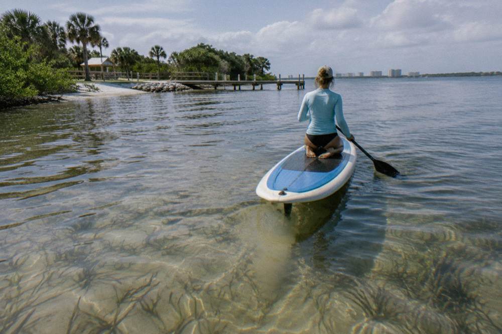 A paddleboarder kneeling on calm shallow lagoon water near seagrass