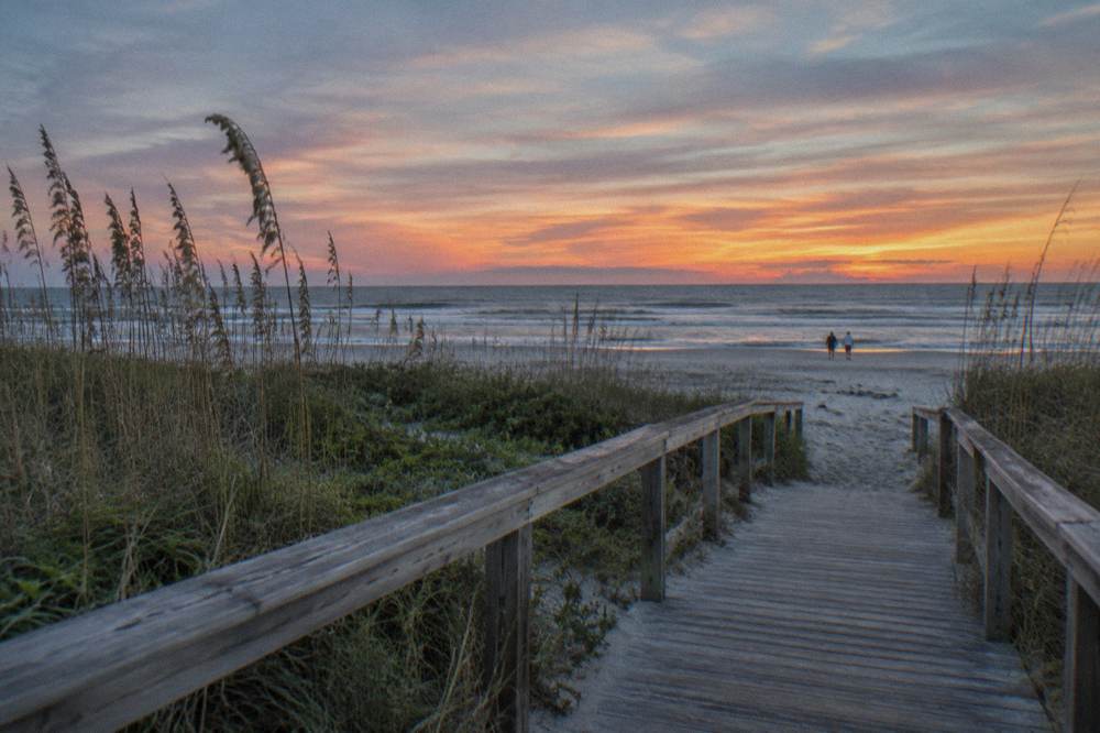 An empty boardwalk over dune grass under a wide pink and orange evening sky