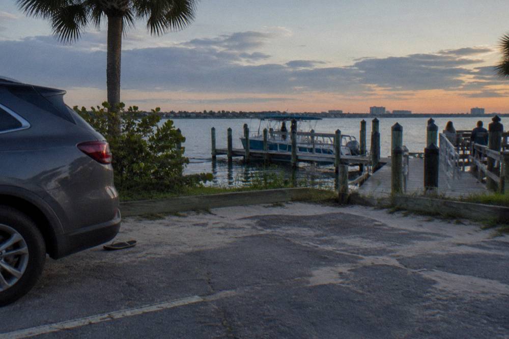 A dusk riverside parking area with parked cars beside calm lagoon water and docks